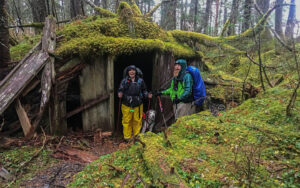 Michelle, Ranger B, and dog Lilly in front of a mossy cabin in Caines Head, Alaska.